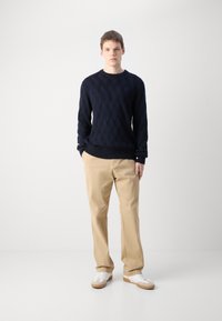 Maglione lavorato a maglia di un blu navy scuro con un motivo a diamante testurizzato, abbinato a pantaloni beige e sneakers bianche con suole marroni.