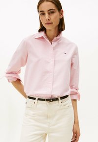 Camisa a rayas rosa claro con cuello abotonado, mangas largas remangadas y un pequeño logo en el pecho izquierdo. Combinada con pantalones blancos.