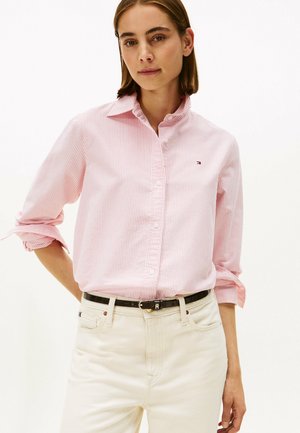 Camisa a rayas rosa claro con cuello abotonado, mangas largas remangadas y un pequeño logo en el pecho izquierdo. Combinada con pantalones blancos.