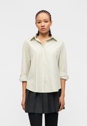 Femme aux cheveux tressés portant une chemise à rayures pâles avec manches retroussées, une jupe plissée foncée et un pantalon noir, debout devant un fond blanc.