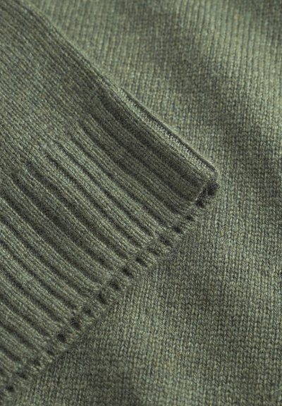 Tissu tricoté vert présentant des poignets côtelés et une texture de corps lisse, mettant en valeur un mélange de fils fins et grossiers dans des teintes variées.