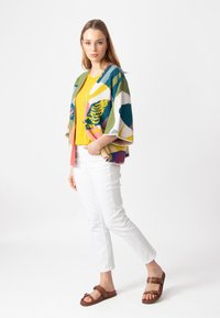 Kleurige gebreide cardigan over een gele top, gecombineerd met witte broek en bruine sandalen. De cardigan heeft gedurfde, abstracte ontwerpen.