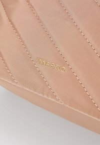 Beige kviltat tyg med diagonala sömmar och guld "ANNA FIELD" märke präglat på ytan.