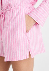 Rosa og hvite stripete shorts med elastisk midje, sidelommer og sidesplitter i kanten. Laget av lett stoff med myk tekstur.