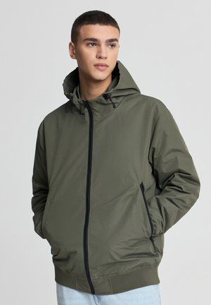 Giacca con cappuccio verde oliva realizzata in tessuto liscio e leggero. Presenta una cerniera frontale, tasche laterali e polsini ed orlo elasticizzati.