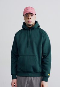 Dark green hooded sweatshirt with a front pocket and adjustable drawstrings, paired with a pale pink cap. Features ribbed cuffs.