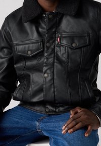 Black leather jacket with a textured finish, featuring a shearling collar, two chest pockets, and silver hardware accents. Worn over blue denim.