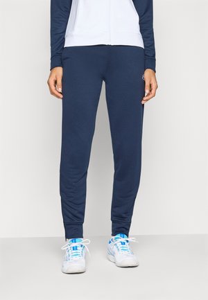 Person som bär marinblå joggerbyxor, vita och blå sneakers samt en vit och marinblå jacka med dragkedja, står framför en enfärgad bakgrund.