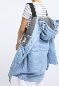 Doudoune bleu clair avec accents gris, boutons-pression et capuche amovible, portée sur une chemise rayée. Tissu doux et texturé.