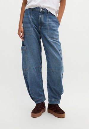 Person trägt lockere blaue Denim-Cargohosen mit großen Taschen und braune Sneakers, steht mit einer Hand in der Tasche.