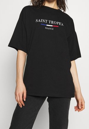 Persona con una camiseta negra holgada con "Saint Tropez France" y una franja azul, blanca y roja en el pecho, combinada con pantalones negros.