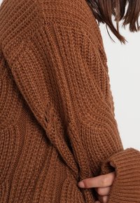 Dzianinowy brązowy sweter z teksturowym wzorem i oversize'owym krojem. Posiada ściągacze na rękawach oraz masywny fason, podkreślający przytulny materiał.