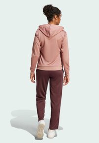Rosa huvtröja med en mjuk textur, i kombination med mörk vinröda sweatpants. Vita sneakers fulländar outfiten. Bakifrån.