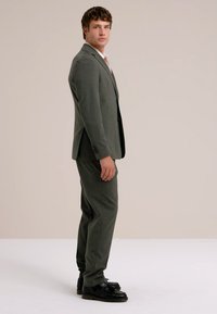 Completo grigio a due pezzi realizzato in tessuto liscia, caratterizzato da una vestibilità su misura, abbinato a scarpe nere e una camicia chiara.