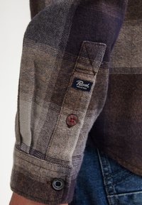 Camicia di flanella a scacchi in tonalità marrone, grigia e navy, con polsini con bottoni, etichetta di marca e tessuto testurizzato.