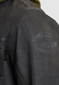 Zwarte leren jas met geborduurd "PME Legend American Classic" logo, gestructureerde afwerking, zichtbare stiksels en een stoffen label.