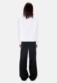 Άτομο με μακριές ραβδώσεις, φορώντας λευκό πουκάμισο με μακριά μανίκια και μαύρο παντελόνι με φαρδιά πόδια, στέκεται με πλάτη γυρισμένη σε απλό φόντο.