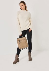 Beige ribbed sweater, black skinny jeans, and brown ankle boots. Carrying a tan textured backpack with dual straps and zipper accents.