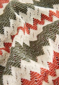 Nahaufnahme eines strukturierten Stoffes mit Zickzackmuster in den Farben Creme, Olivgrün und Rostrot, mit sichtbaren Nähten und Falten.