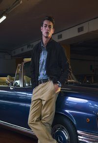 Man in black jacket, denim shirt, and beige pants leans against a blue vintage convertible car in a dimly lit indoor garage.