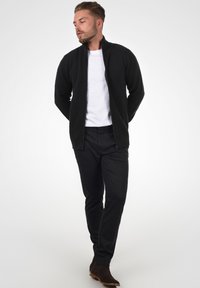 Schwarze gerippte Zip-Jacke mit hohem Kragen, getragen über einem weißen T-Shirt. Kombiniert mit strukturierten schwarzen Hosen und dunkelbraunen Stiefeletten.