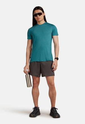 Teal t-shirt med kort ärm och grå shorts, med svarta sportskor. Silverfärgad vattenflaska i rostfritt stål och stora solglasögon.