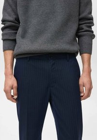 Pulover gri tricotat cu model reliefat, asortat cu pantaloni bleumarin cu dungi subțiri, având un front drept și o închidere cu nasture.