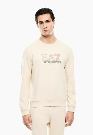 Młody mężczyzna o ciemnych włosach, ubrany w kremową bluzę i spodnie z logo „EA7 Emporio Armani” na piersi, stojący na białym tle.
