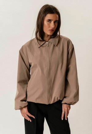 Jauna moteris dėvi smėlio spalvos užtraukiamą džemperį su apykakle ir juodus kelnes, stovinti priešais paprastą šviesų foną.