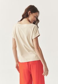 Top bej cu mânecă scurtă, cu o croială lejeră și decolteu la spate, asociat cu pantaloni roșii cu un croi fluid. Textură netedă, design minimalist.