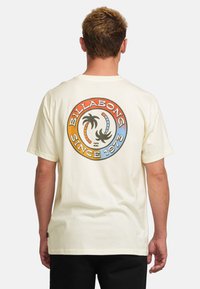 Cream short-sleeve t-shirt featuring a colorful circular logo with palm trees and “BILLABONG SINCE 1973” text on the back.