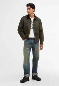 Casaco verde-oliva com um colarinho de cortiça castanho, fecho frontal de botões e quatro bolsos, combinado com jeans azuis desbotados e sapatos pretos.