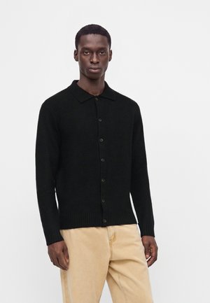 Homme portant un cardigan noir boutonné avec col et un pantalon beige sur un fond blanc uni.