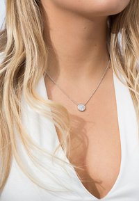Collana d'argento con un pendente rotondo, caratterizzato da una superficie testurizzata, sospesa a una delicata catena. Indossata sopra un top bianco a scollo a V.