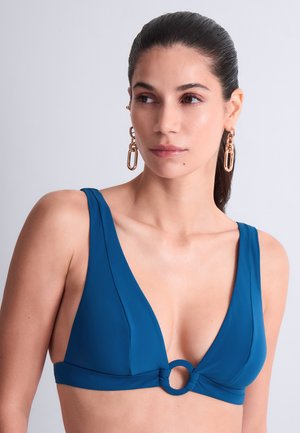 Femme aux cheveux foncés tirés en arrière, portant des boucles d’oreilles en chaîne dorée et un haut de bikini bleu à décolleté profond en V avec un anneau central.