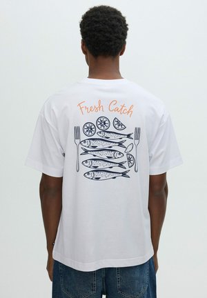Persona con camiseta blanca con texto "Fresh Catch", pez ilustrado, rodajas de limón y dos tenedores en la espalda.