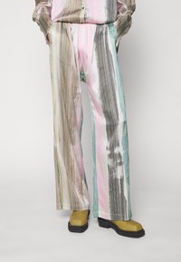 Pestrobarevné kalhoty s širokými nohavicemi, zdobené vertikálními pruhy v růžové, hnědé a tyrkysové barvě. Materiál vypadá hladce s lesklým povrchem. Černé chunky boty.