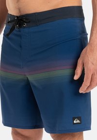 Shorts de bain bleu marine avec un motif à rayures horizontales en rouge, vert et jaune. Dotés d'une taille noire avec cordon de serrage et d'un patch logo sur le côté.