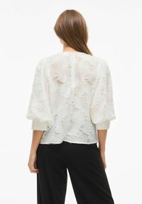 Blouse blanche texturée avec des manches bouffantes trois-quarts, présentant un design superposé transparent et des détails à bords bruts, portée sur un pantalon noir.