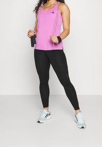 Top deportivo rosa con tirantes finos, leggings negros y zapatillas de correr azul claro. Sosteniendo una cuerda para saltar. Los leggings tienen detalles laterales.
