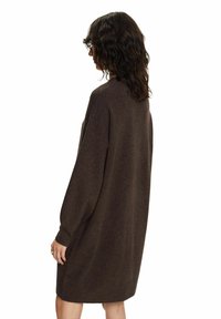 Femme aux cheveux bouclés mi-longs portant une robe-pull ample à manches longues, de couleur brun foncé, arrivant au niveau des genoux, debout de profil sur fond blanc.
