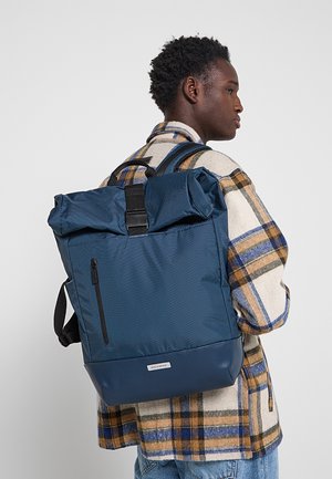 Uomo con giacca a quadri che indossa un grande zaino blu con chiusura roll-top, tasca frontale con cerniera ed etichetta del brand, di profilo.