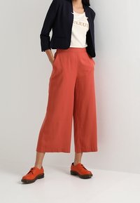 Femme portant un pantalon large taille haute couleur rouille, des chaussures lacées orange, un tee-shirt blanc à motif et un blazer court bleu marine, debout contre un mur blanc.