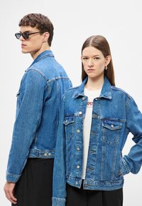 Világoskék farmerdzsekik gombos záródással, mellzsebekkel és kontrasztos varrással. Az egyik nyitva van viselve; a másik zsebbe tett kézzel.