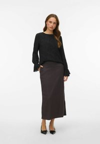 Melna garām piedurknēm krekls ar fakturētu rakstu un izplatītiem aproču galiem, kopā ar tumšu svītrainu maxi svārku. Valkāti ar melnām smailām kurpēm.