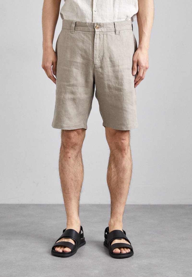 Shorts en lin beige avec une coupe décontractée, dotés d'un devant plat, d'une fermeture à bouton et de détails cousus. Associés à des sandales noires à enfiler.