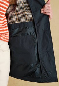 Dłoń trzymająca otwartą czarną kurtkę, ukazującą wnętrze z dużą kieszenią i wzorem w kratę, na ręku pomarańczowo-biały sweter w paski.