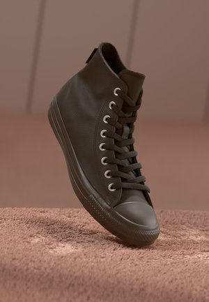 Ciemnobrązowy sneaker za kostkę z sznurowadłami, stojący pionowo na teksturowanej brązowej powierzchni na jednolitym brązowym tle.