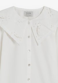 Blusa blanca con un gran collar bordado que presenta intrincados patrones recortados. Abotonada con botones transparentes en la parte delantera.