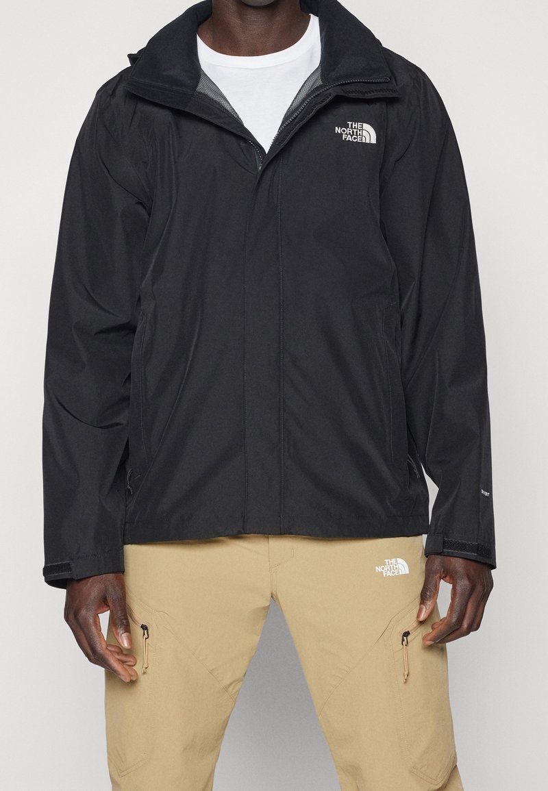 Veste imperméable noire avec fermeture éclair à l'avant, col réglable et détail de logo. Présente une texture lisse et une coupe décontractée.